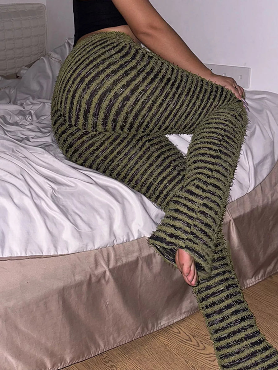 A person sitting on a bed wearing chunky olive green and black striped fuzzy knit pants with open toes.