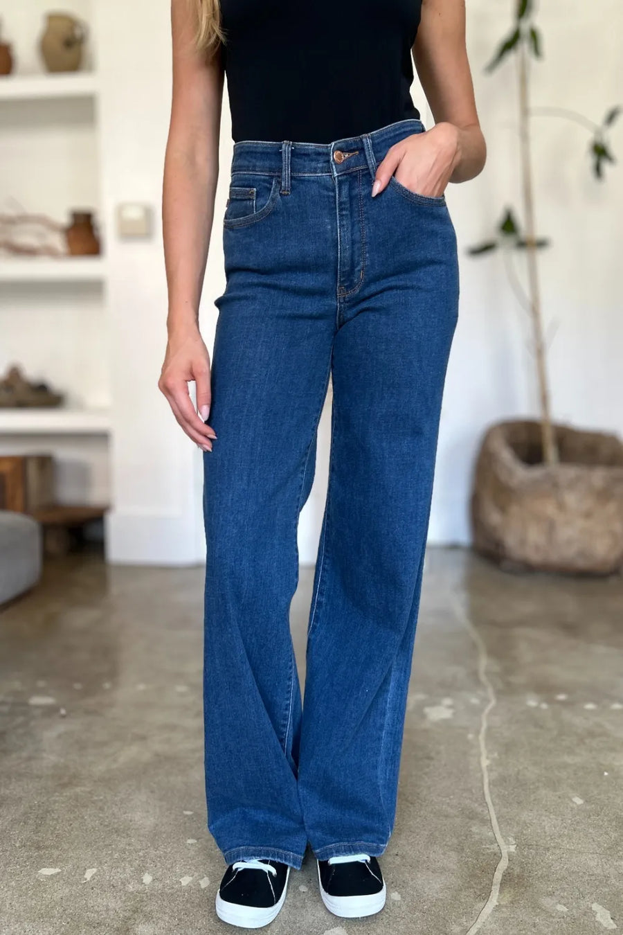 Person wearing blue jeans and black sneakers in a room with a white wall and plant.