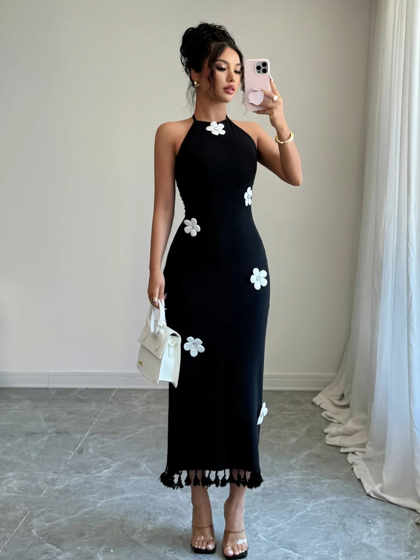 Woman wearing a black halter neck dress with white floral patterns, standing in a room with light-colored curtains.
