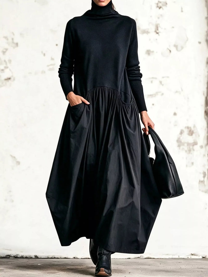 A model wearing a long-sleeve black turtleneck dress with side pockets, paired with black ankle boots and carrying a black tote bag, set against a textured white wall.