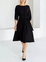 A woman modeling a black midi dress with three-quarter sleeves, a boat neckline, and a matching fabric tie belt at the waist, paired with black pointed-toe heels in a bright white studio setting.