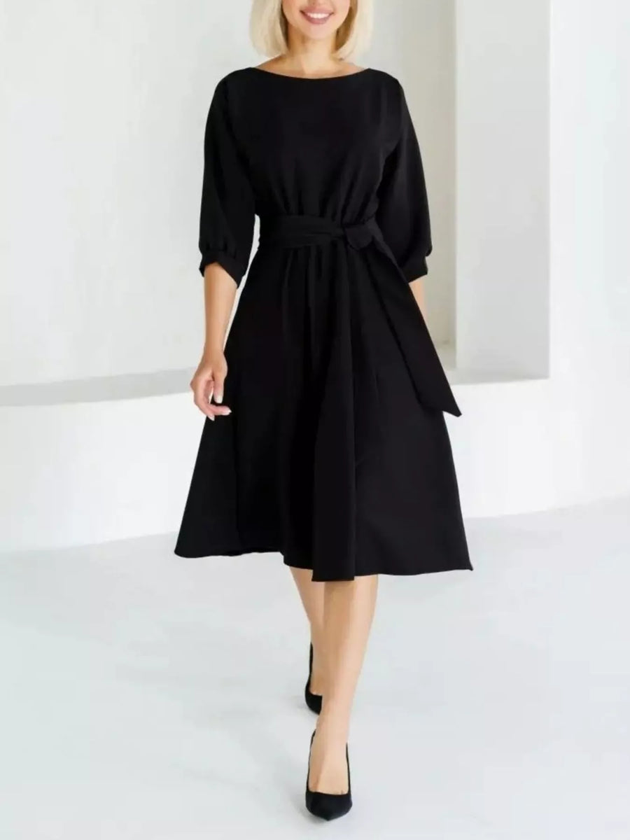 A woman modeling a black midi dress with three-quarter sleeves, a boat neckline, and a matching fabric tie belt at the waist, paired with black pointed-toe heels in a bright white studio setting.