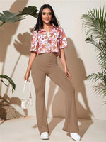 Woman wearing a floral blouse and brown pants standing against a neutral background with plants.