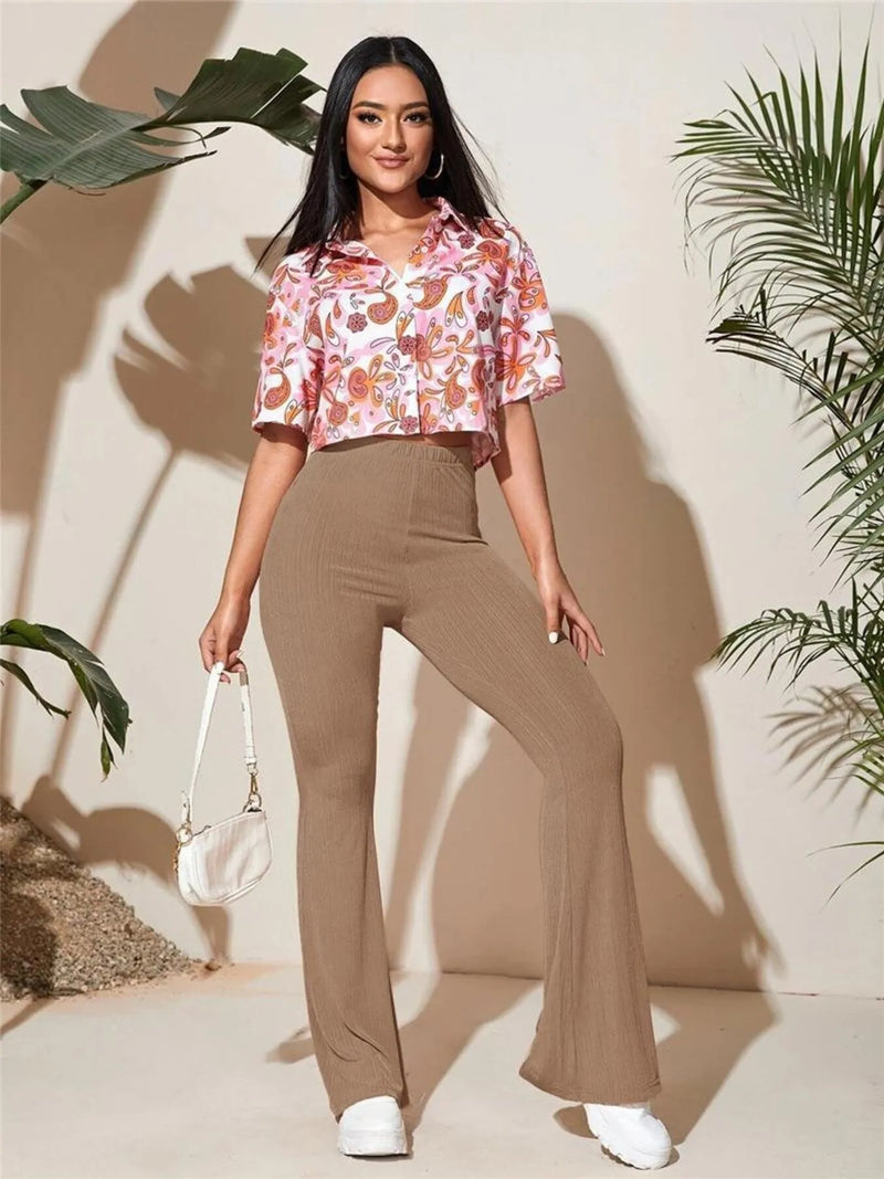 Woman wearing a floral blouse and brown pants standing against a neutral background with plants.