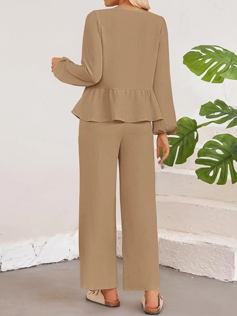 Person wearing a beige peplum top and wide-leg pants against a white wall with green leaves.