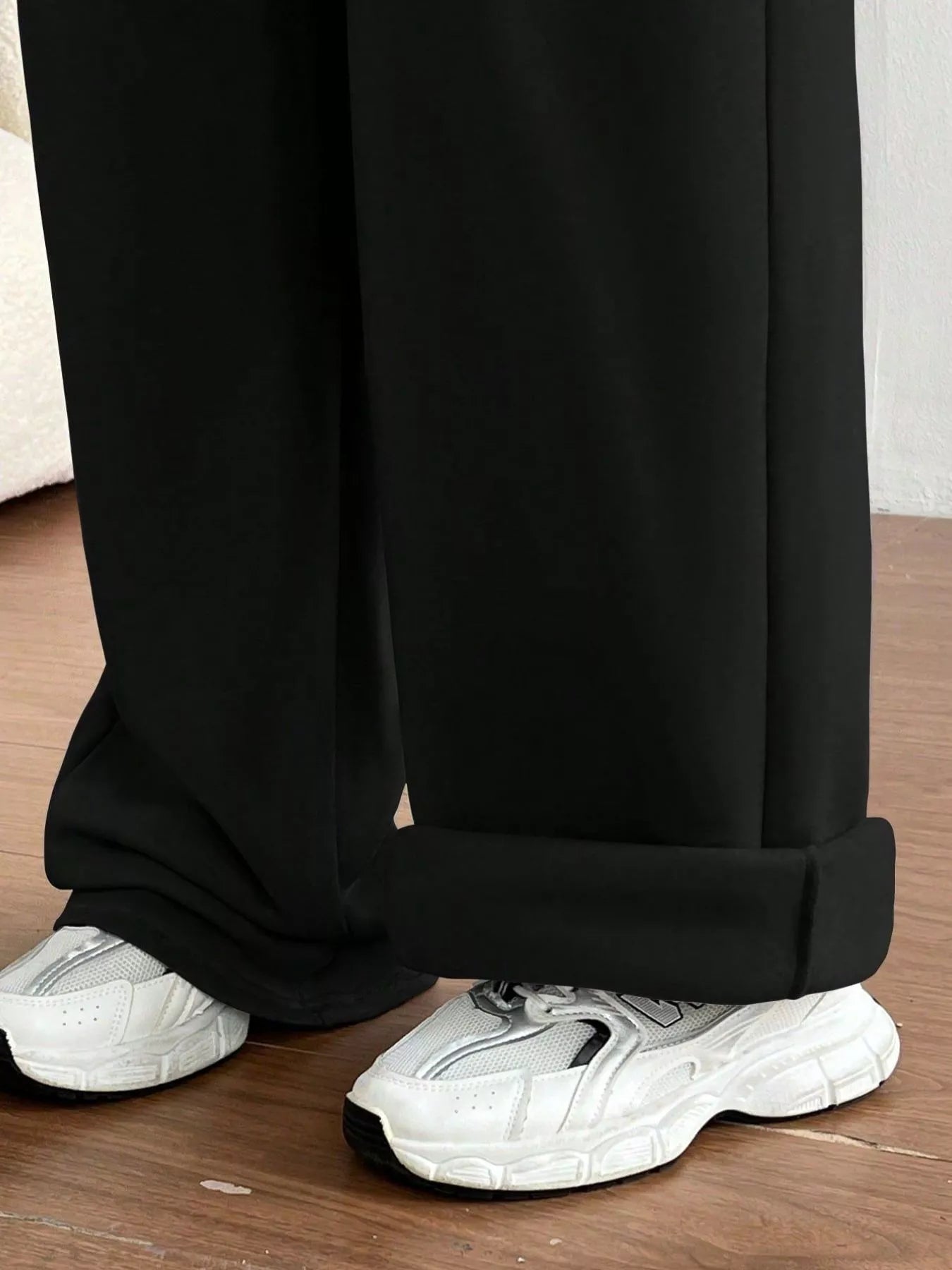 Person wearing black pants with a cuffed hem and white sneakers on a wooden floor.