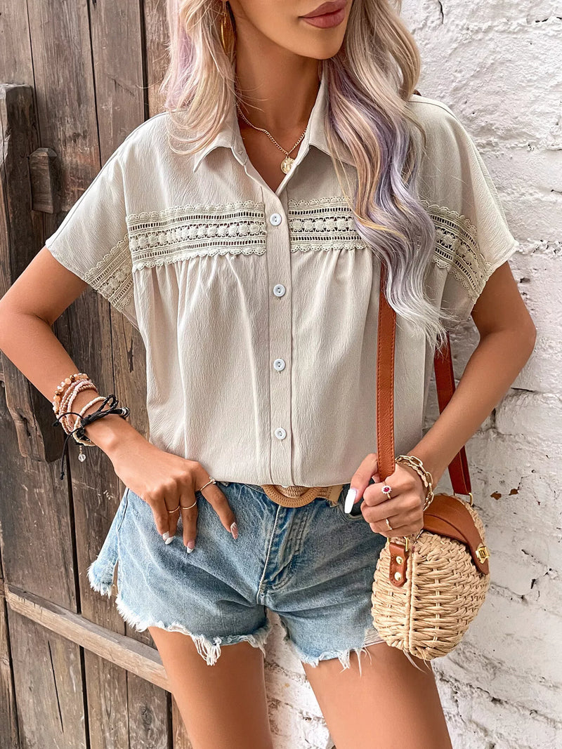 Woman wearing a light-colored blouse with lace details and denim shorts, standing against a rustic wooden wall.