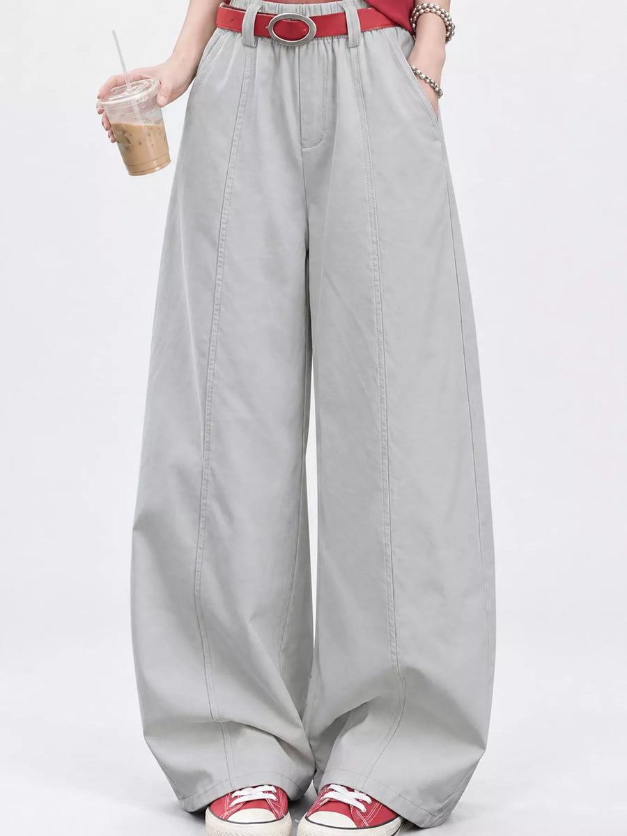 A person wearing light gray wide-leg pants with a red belt, holding an iced coffee, and wearing red and white sneakers against a plain white studio background.