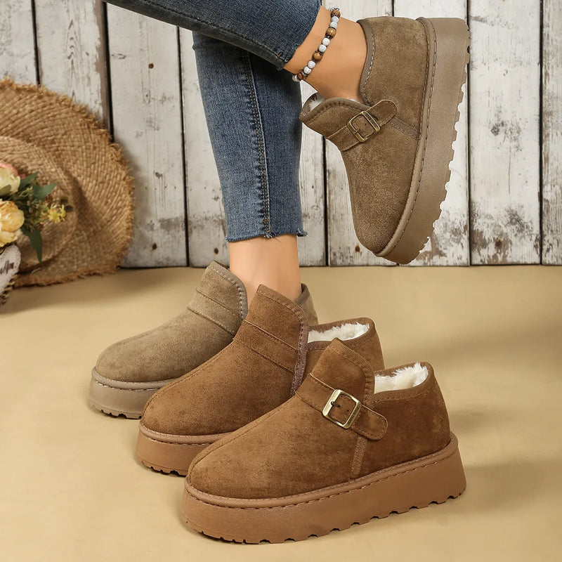 Brown suede boots with buckle details worn by a person on a wooden floor.