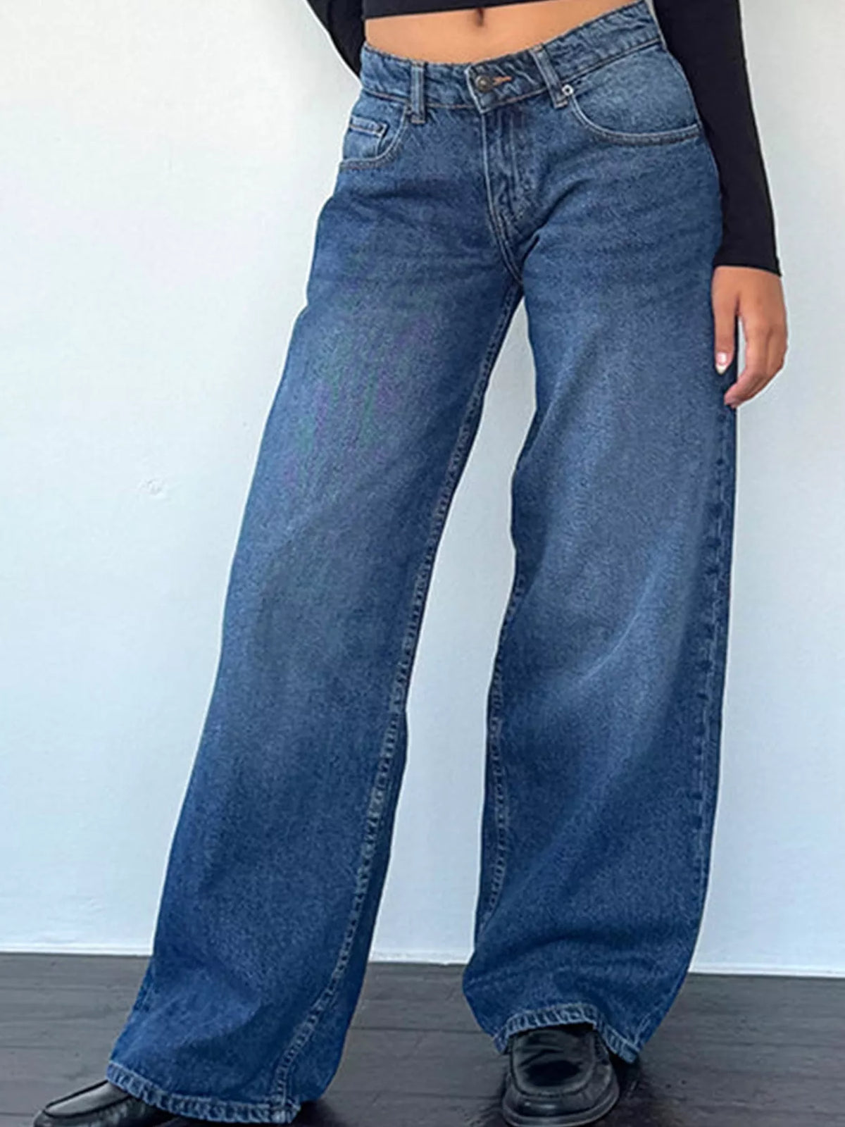 A person wearing high-waisted, wide-leg blue denim jeans with a black long-sleeve crop top and black shoes, standing against a plain white wall.