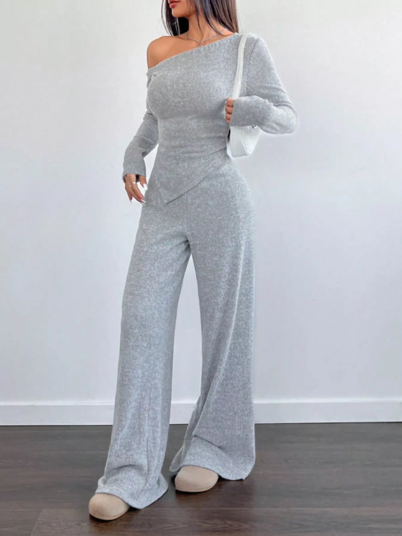 Woman wearing a gray off-shoulder jumpsuit against a white wall.