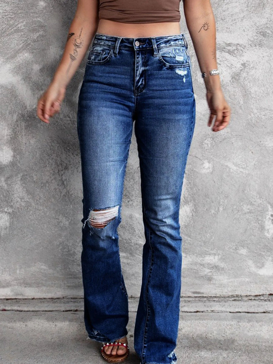 Person wearing blue flared jeans with a brown top against a gray concrete wall.