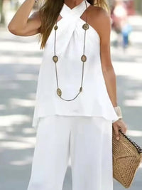 Woman wearing a white sleeveless top and pants with a long necklace and holding a woven clutch.