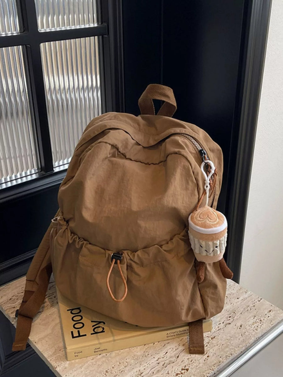 Brown backpack with a small plush toy on top, placed on a surface near a window.