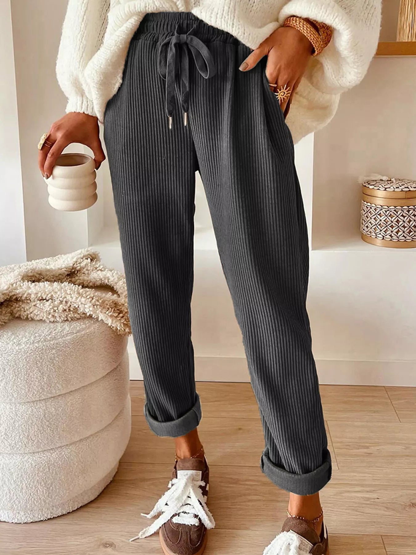 Person wearing dark ribbed pants with a white sweater and brown shoes, holding a ceramic cup.