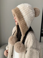 Person wearing a fluffy, two-tone hat with pom-poms indoors.