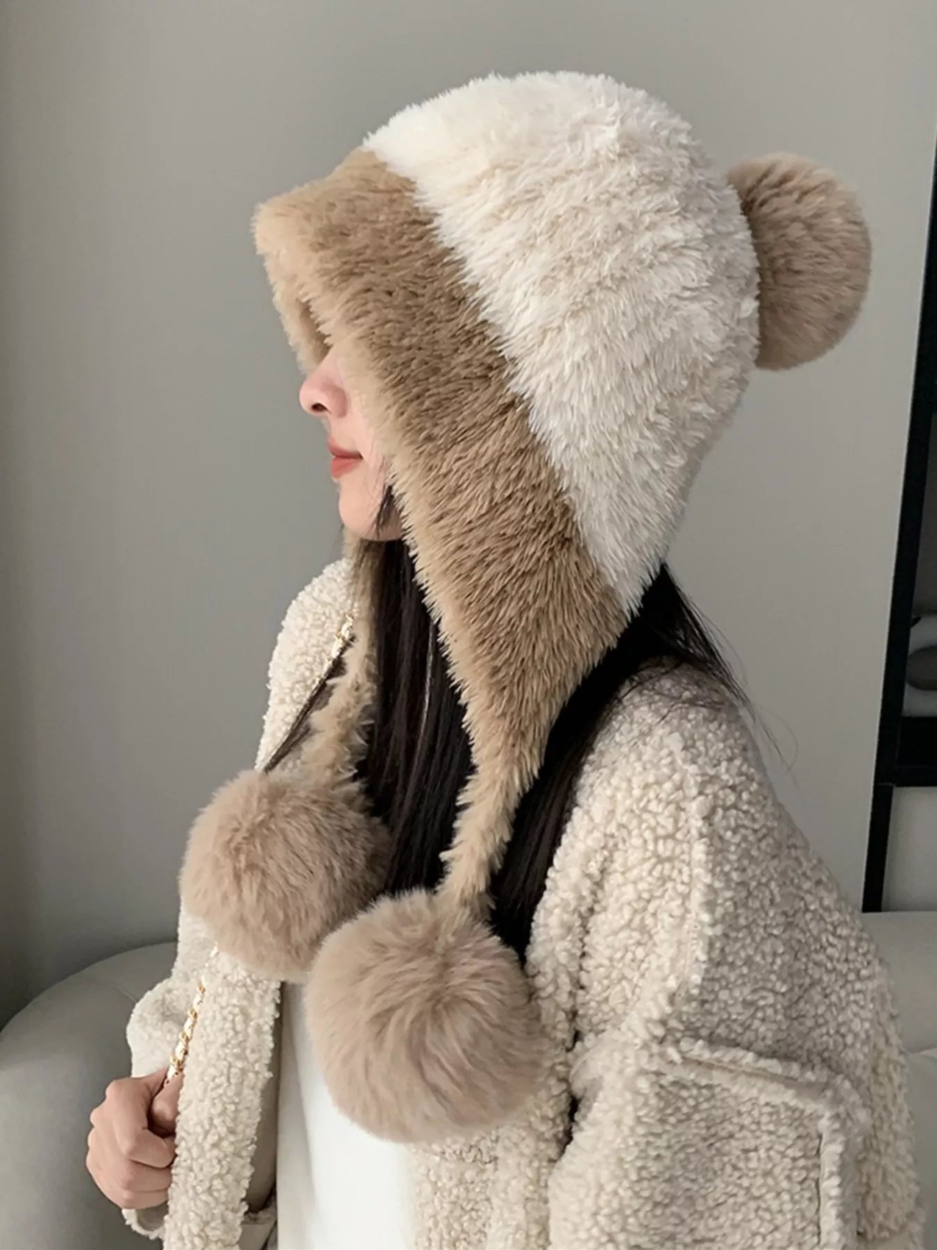 Person wearing a fluffy, two-tone hat with pom-poms indoors.