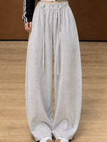 A person wearing light gray wide-leg sweatpants with a drawstring waist, shown from the waist down against a brown wall and wood floor.