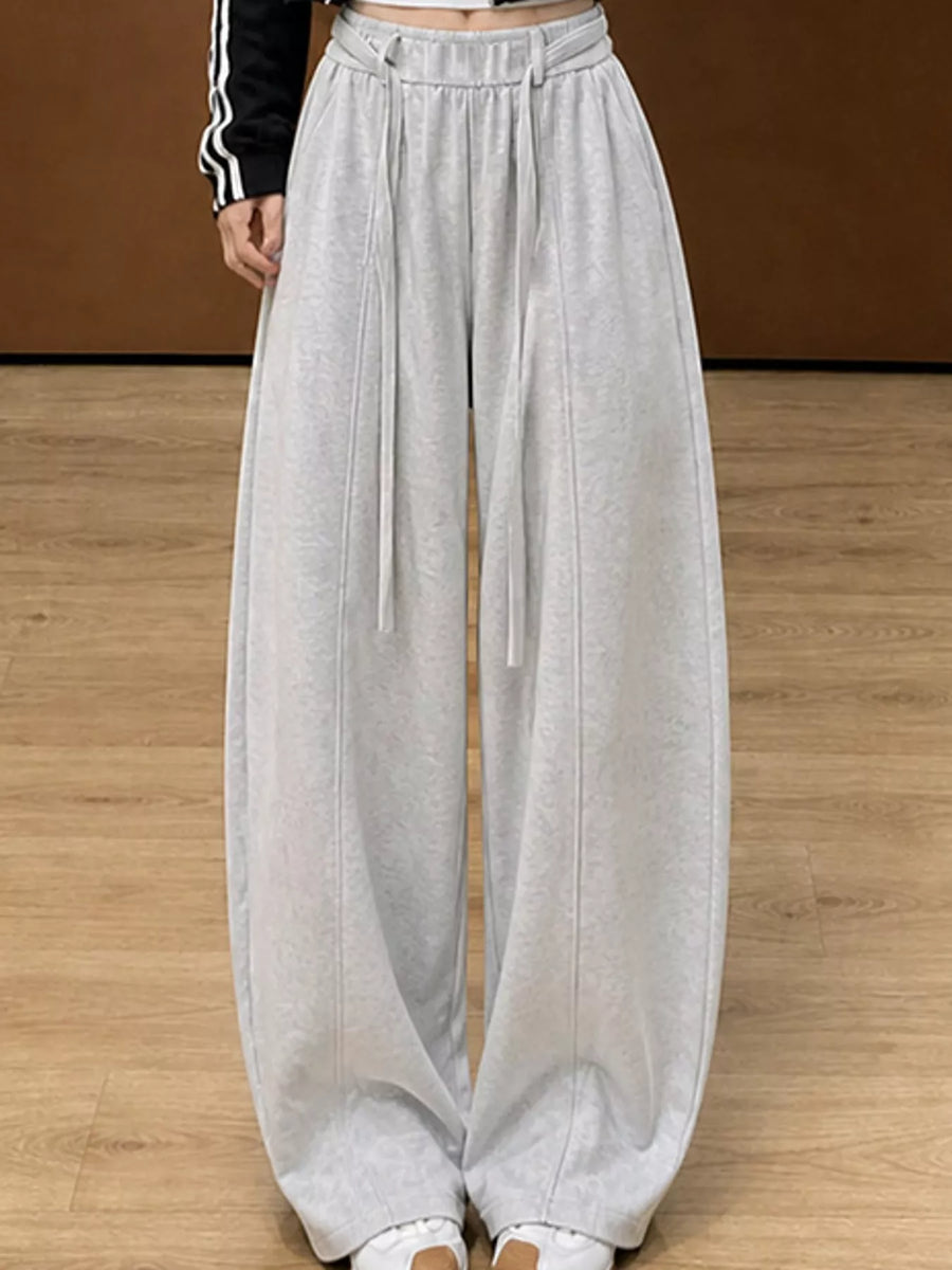 A person wearing light gray wide-leg sweatpants with a drawstring waist, shown from the waist down against a brown wall and wood floor.