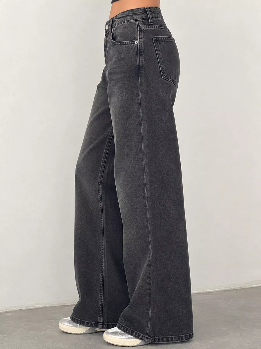 A person wearing dark gray wide-leg jeans, shown from the waist down against a plain light gray wall and concrete floor.