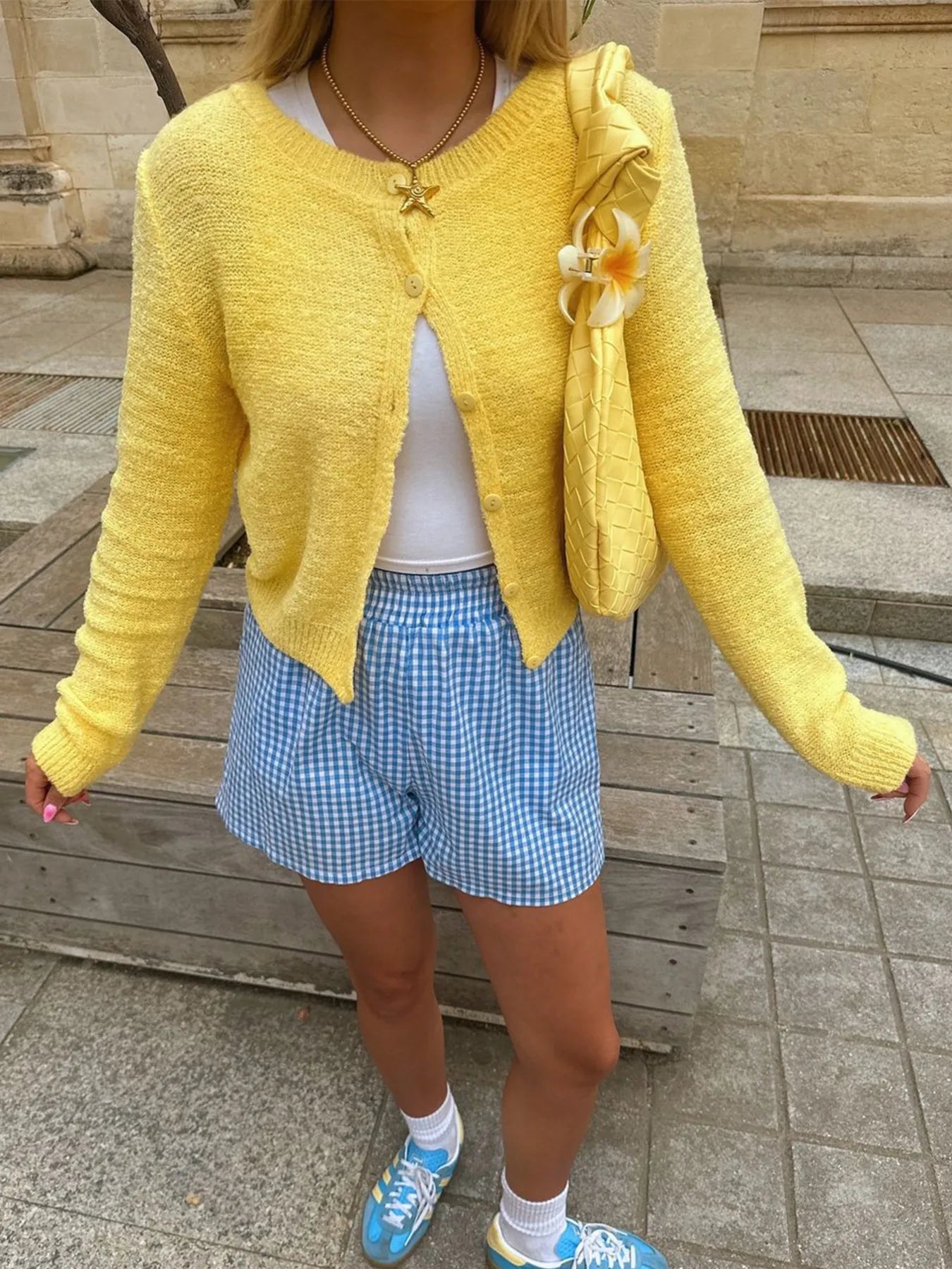 Person wearing a yellow cardigan, white top, blue checkered shorts, and blue sneakers on a stone pavement.