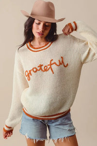 Woman wearing a cream sweater with 'grateful' text and a beige hat, standing against a plain background.