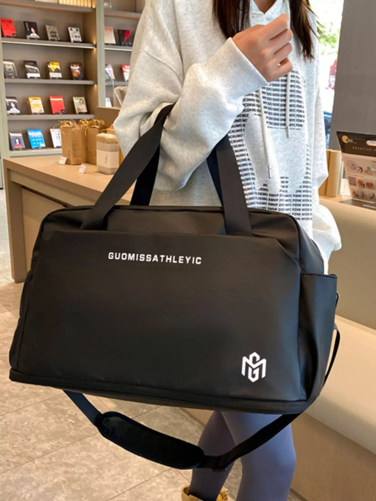 Person holding a black duffel bag with 'GUOMISSATHLETIC' branding in an indoor setting.