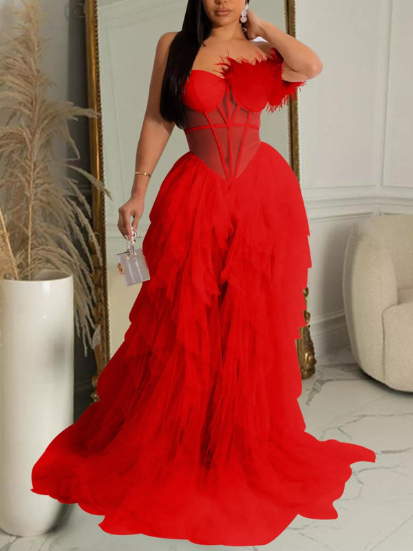 A woman wearing a dramatic bright red formal gown with a structured sweetheart bodice, sheer mesh corset-style panels, feathered shoulder details, and a voluminous layered tulle skirt, standing indoors in a decorated room.