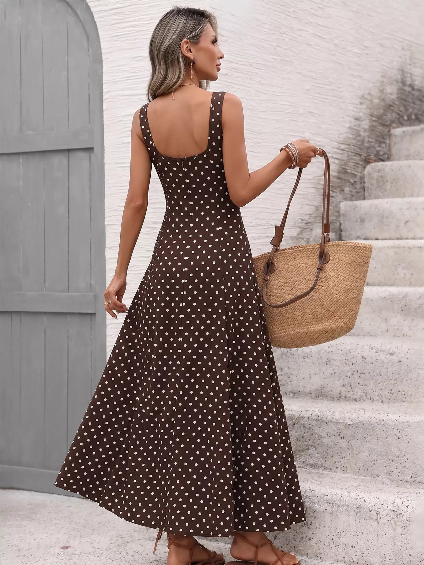 Woman in a brown polka dot dress holding a straw bag on stone steps.