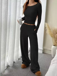 Person wearing a black ribbed outfit with wide-leg pants in a room with white curtains and a plant.