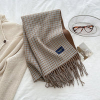Plaid scarf with tassels on a white surface, next to a beige cardigan and glasses.