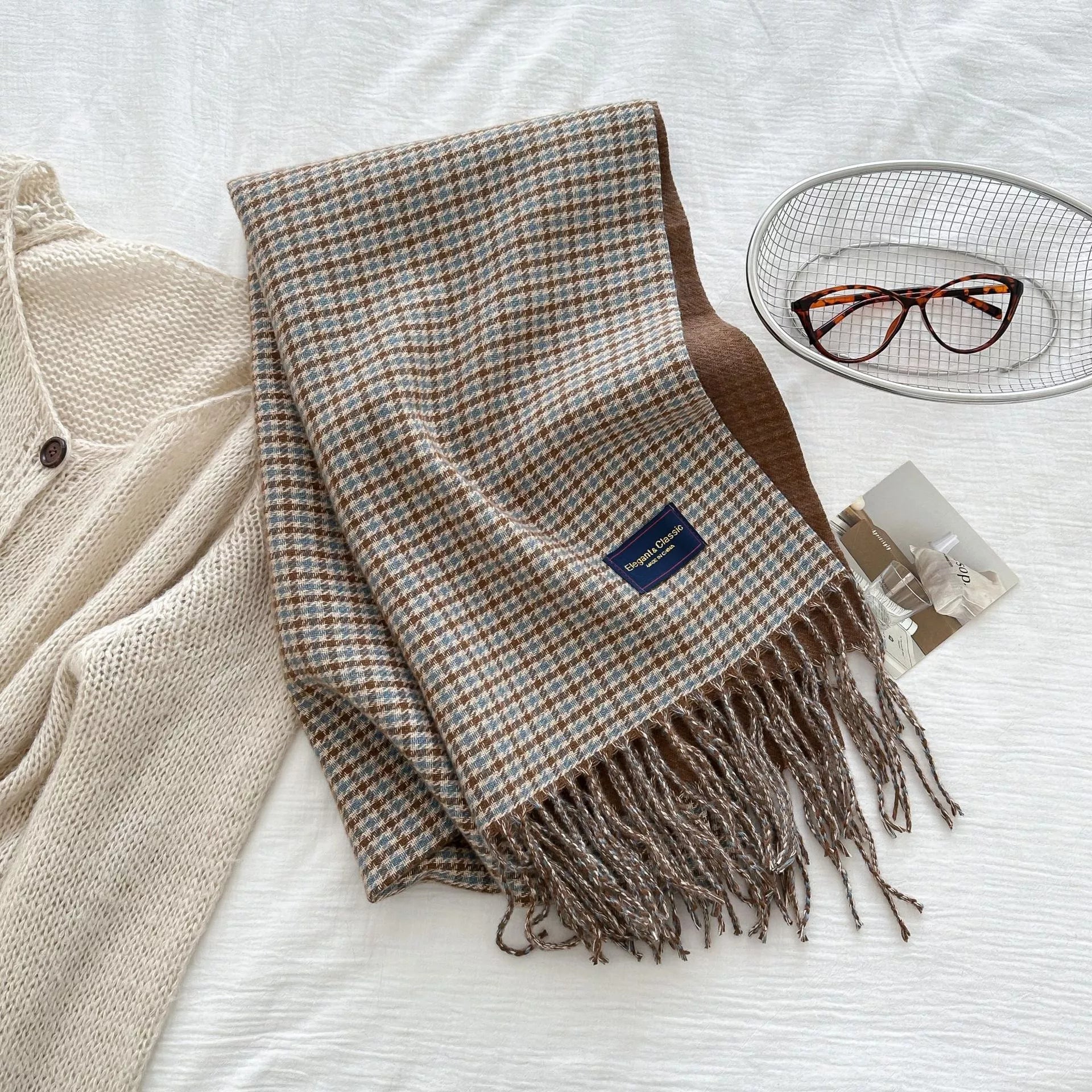 Plaid scarf with tassels on a white surface, next to a beige cardigan and glasses.