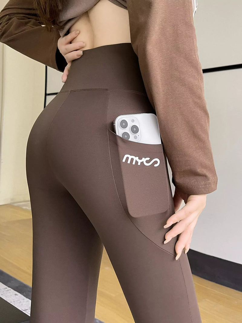 A close-up of a person wearing high-waisted brown leggings with a side pocket holding a white smartphone. The pocket has a white 'mymo' logo, and the person is also wearing a matching brown long-sleeve top.