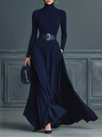 Woman wearing a long navy dress with a belt in a room with gray walls.