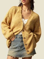 A woman wearing a fuzzy mustard yellow open-front cardigan with scalloped trim and brown buttons, layered over a white top and paired with a denim mini skirt, against a solid light beige background.