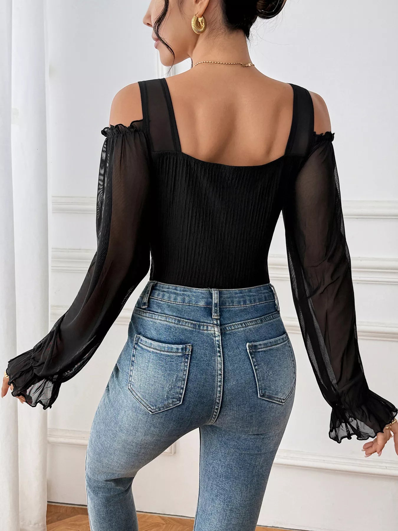 Person wearing a black top with sheer sleeves and blue jeans in a neutral setting