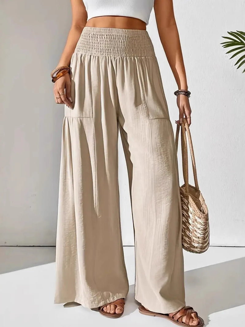 Person wearing beige wide-leg pants and a white top, holding a woven bag on a plain background