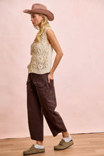 Woman wearing a beige sleeveless top, brown wide-leg pants, and a wide-brimmed hat against a pink background.