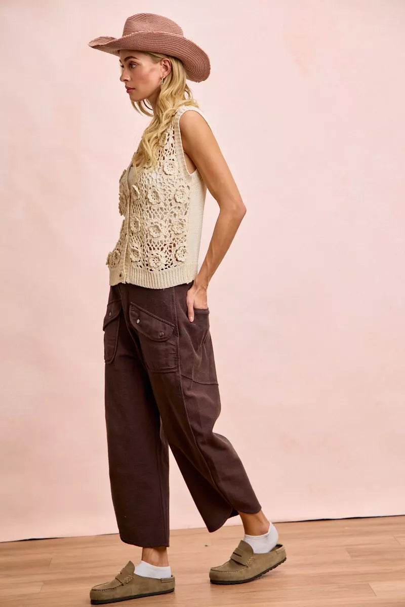 Woman wearing a beige sleeveless top, brown wide-leg pants, and a wide-brimmed hat against a pink background.