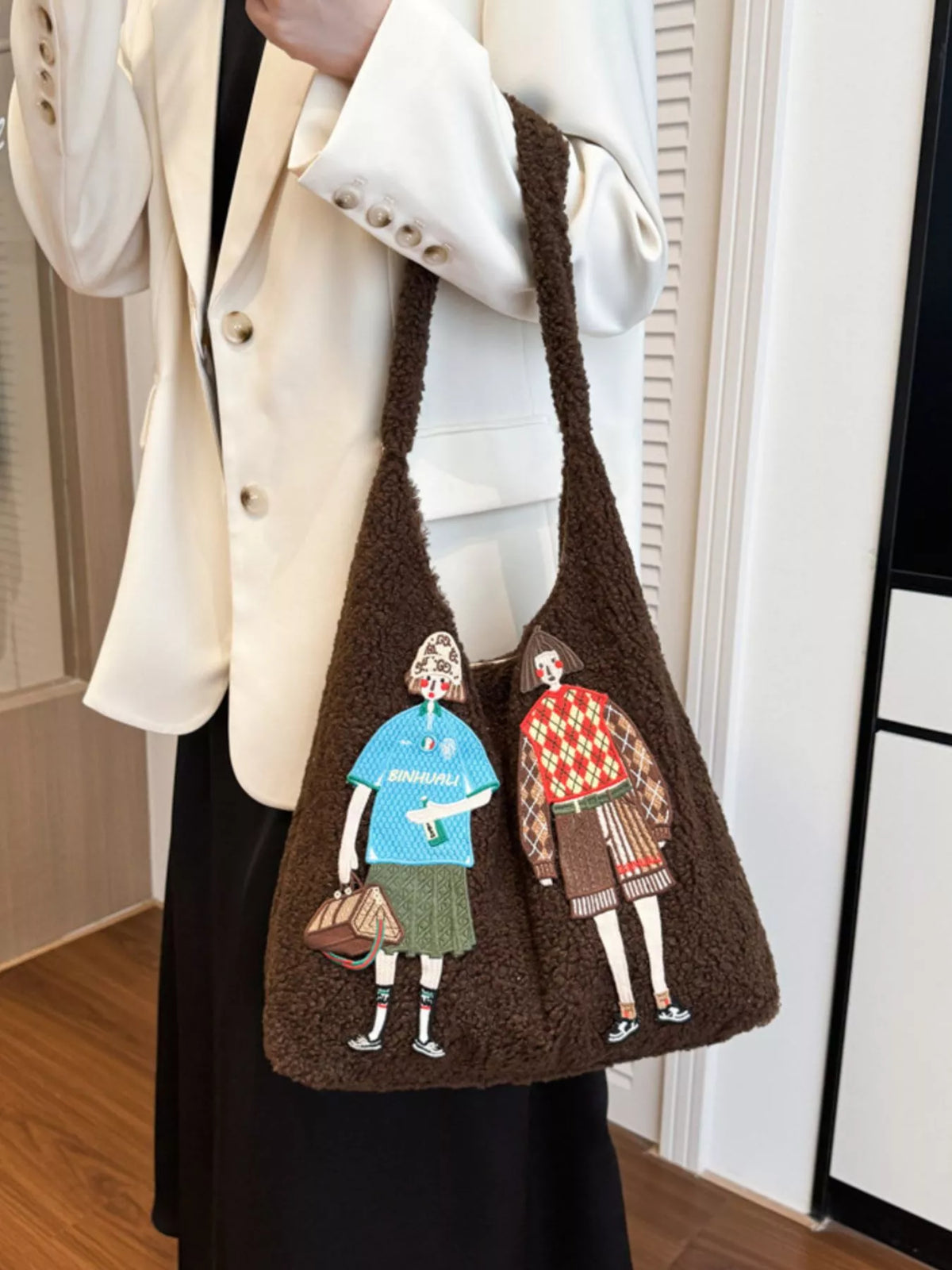 Brown tote bag with embroidered figures held by a person wearing a white coat.