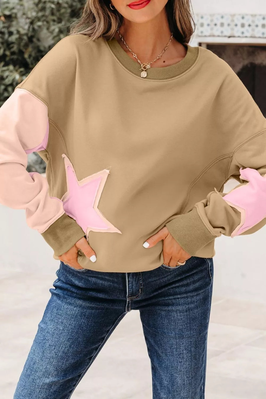 A woman wearing a beige crewneck sweatshirt with light pink star-shaped patches on the front and light pink elbow patches, paired with blue jeans.