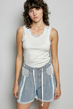 Woman wearing a white sleeveless top with lace trim and denim shorts on a plain background