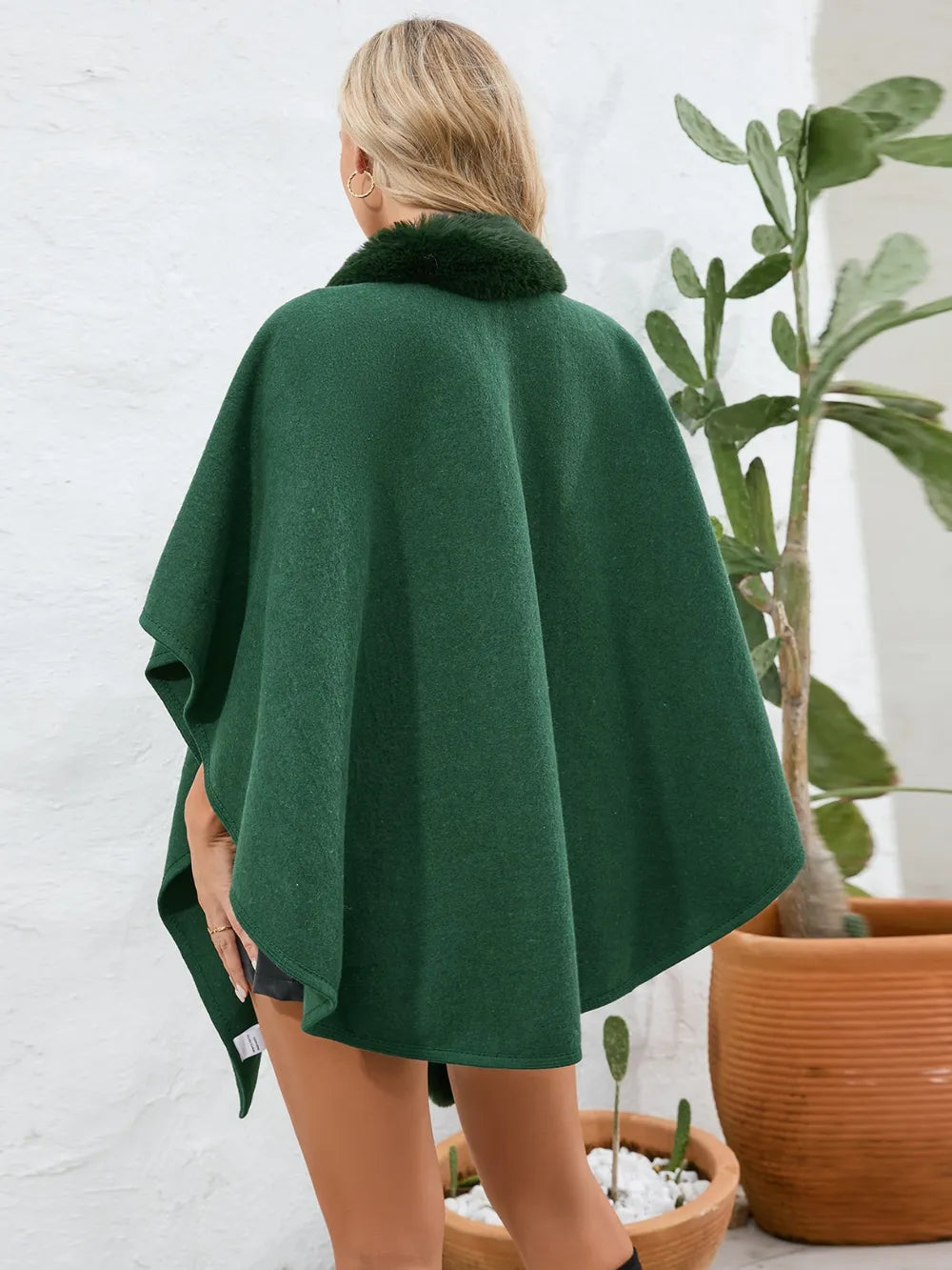 Person wearing a green poncho standing in front of a white wall with plants.