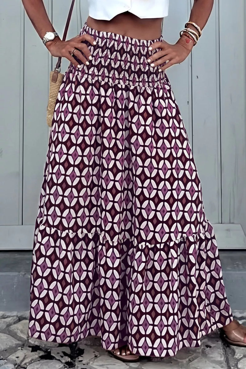Person wearing a long patterned skirt with a white top and sandals, standing against a light-colored wall.