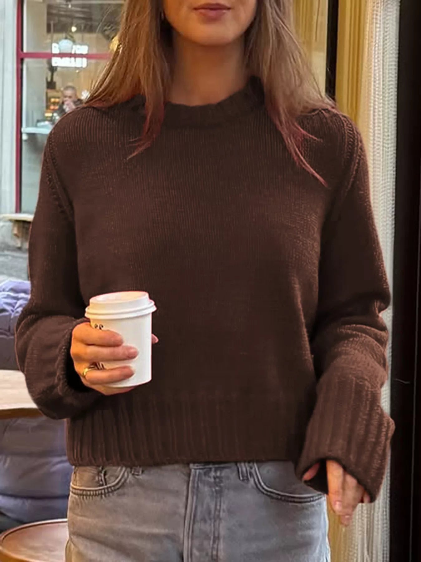 Person wearing a brown sweater holding a white coffee cup indoors.