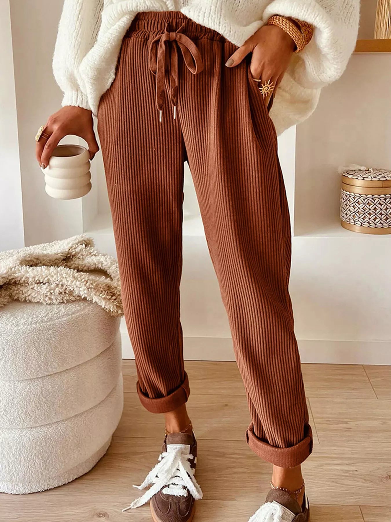 Person wearing brown ribbed pants and white sneakers, holding a ceramic cup indoors.