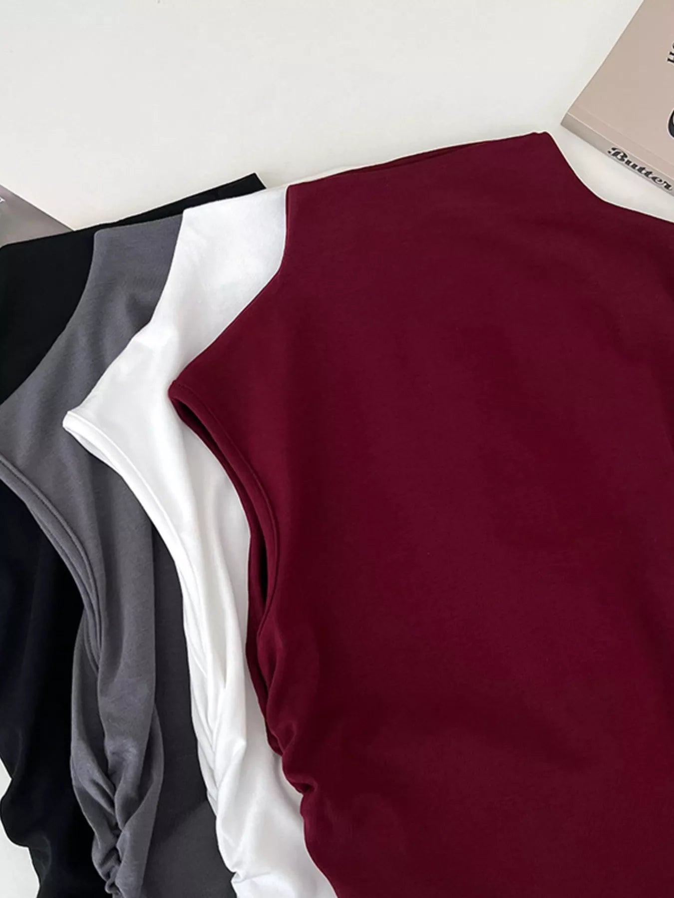 Three sleeveless tops in black, white, and burgundy on a light gray background.