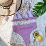 Purple bikini set on a white lace surface with a hat, avocado, and lemon.