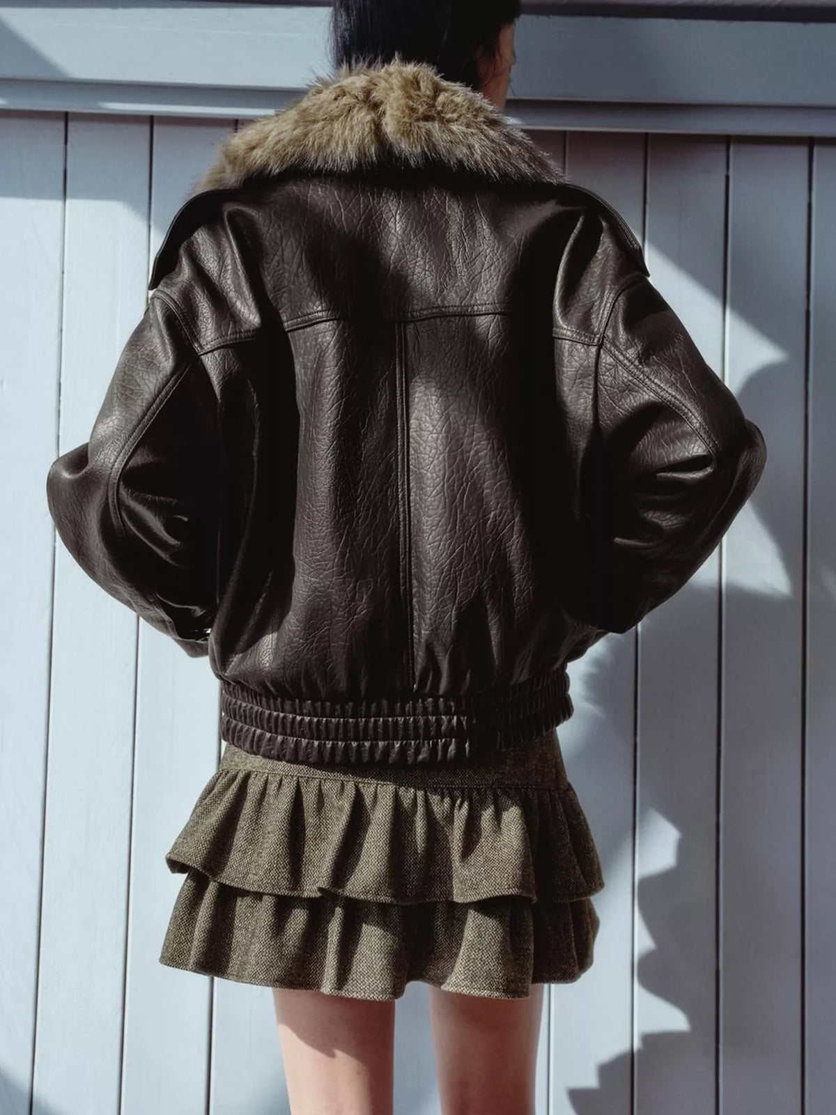 A person shown from the back wearing a dark brown leather jacket with a tan faux-fur collar and an olive green ruffled mini skirt, standing against a white paneled wall.
