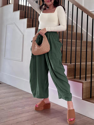 Woman in green pants and white top holding a woven handbag on a wooden floor.